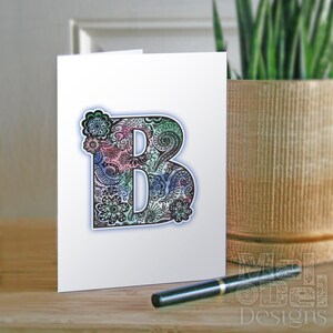 May include: A white greeting card featuring a large, ornate letter "B" with a floral and paisley design in shades of pink, blue, and green. A black pen rests on the wooden surface in front of the card. The card is next to a potted plant.