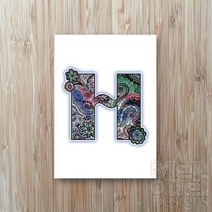 May include: A white card featuring the letter "H" with a floral design. The letter is filled with intricate patterns in shades of blue, pink, and green. Floral accents are placed around the letter. The card is on a wooden surface.