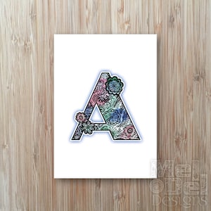 May include: A white card with a black outlined letter A filled with a colorful floral pattern. The letter A is outlined in blue and has a light blue glow. The floral pattern is in shades of pink, blue, and green.