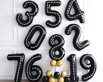 May include: Black and white number balloons, from 0 to 9, with white stitching. The balloons are arranged in a semi-circle on a white wall. There are also black and gold balloons in the bottom center of the image.