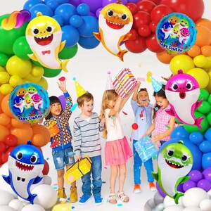 K&ouml;nnte beinhalten: Eine festliche Szene mit feiernden Kindern, umgeben von bunten Luftballons. Haif&ouml;rmige Folienballons und runde Ballons mit dem Text "Baby Shark" sind prominent. Kinder halten Geschenke und tragen Partyh&uuml;te.