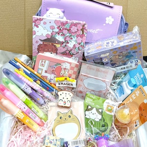 Puede incluir: Una caja morada llena de una variedad de art&iacute;culos de papeler&iacute;a, que incluyen bol&iacute;grafos, l&aacute;pices, borradores, pegatinas y un peque&ntilde;o bolso. La caja est&aacute; llena de papel triturado y los art&iacute;culos est&aacute;n dispuestos de forma visualmente atractiva.