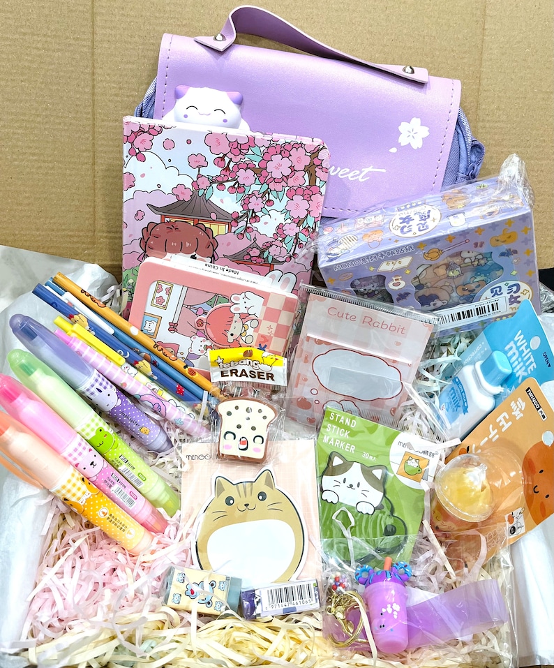 Puede incluir: Una caja morada llena de una variedad de art&iacute;culos de papeler&iacute;a, que incluyen bol&iacute;grafos, l&aacute;pices, borradores, pegatinas y un peque&ntilde;o bolso. La caja est&aacute; llena de papel triturado y los art&iacute;culos est&aacute;n dispuestos de forma visualmente atractiva.