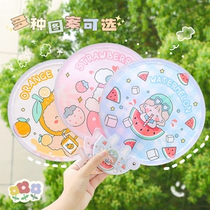 May include: Three round hand fans with different fruit designs. The fans are made of plastic and have a handle. The designs are orange, strawberry, and watermelon. The text on the fans reads "Orange", "Strawberry", and "Watermelon".