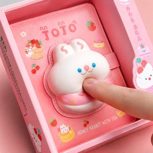May include: A pink notebook featuring a squishy white rabbit face with a donut design on the front cover. The cover includes illustrations of strawberries, cherries, and cakes, with the text JOJO and HONEY RABBIT WITH YOU printed on the surface.