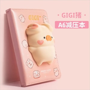 May include: A pink A6 notebook with a 3D pig design. The notebook has a white and pink pig pattern and the text "GIGI" and "SOFT LIFE" on the cover.