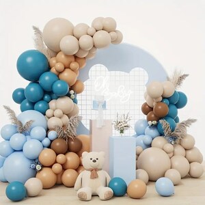 Pode incluir: Um arco de bal&otilde;es para ch&aacute; de beb&ecirc; com bal&otilde;es azuis, marrons e bege. O arco &eacute; decorado com capim-dos-pampas e um urso de pel&uacute;cia. Uma placa de neon diz "Oh Baby".