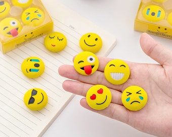 May include: A set of yellow emoji erasers with different facial expressions, including smiling, crying, winking, and sunglasses. The erasers are in a variety of packages and are being held in a hand.