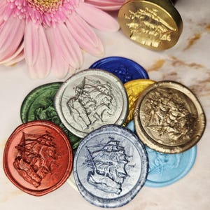 May include: A selection of colourful wax seals featuring a ship design, in shades of red, green, silver, gold, blue, and yellow. A gold seal stamp with a wooden handle is also present, with a pink flower.