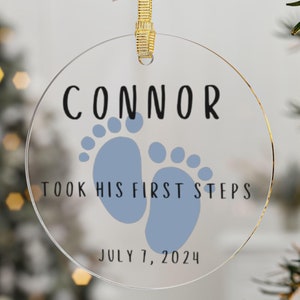 May include: A clear, round ornament with the name "CONNOR" in black lettering. Below are light blue footprints and the text "TOOK HIS FIRST STEPS" and "JULY 7, 2024". The ornament hangs from a gold ribbon.