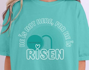 Könnte beinhalten: Tealblaues T-Shirt mit einer weißen Grafik, auf der "Er ist nicht hier, denn er ist auferstanden" steht, mit einem Herz und einer Grabbildgrafik. Matthäus 28,6 ist ebenfalls in der Grafik enthalten.