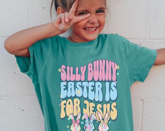 May include: A teal blue t-shirt with a colorful graphic that says "Silly Bunny Easter is for Jesus". Three cartoon bunnies with bows are featured on the graphic.