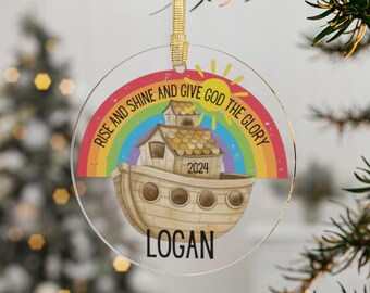 May include: A clear, round Christmas ornament with a gold ribbon. The ornament features a colorful rainbow, a sun, and a depiction of Noah's Ark. Text includes "RISE AND SHINE AND GIVE GOD THE GLORY", "2024", and the name "LOGAN".