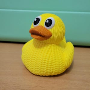 May include: A yellow rubber duck with a textured body and an orange beak. The duck has large, round eyes with black pupils and white highlights. The duck is sitting on a wooden surface.