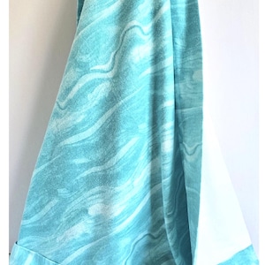 May include: A turquoise blue beach towel with a white border and a white and blue wave pattern. The towel has a small white tag with a blue dragonfly on it.