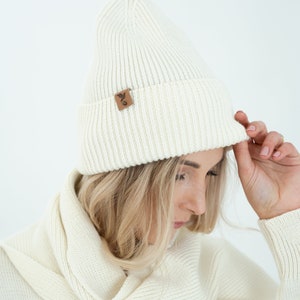 May include: A white knitted beanie with a small brown leather patch featuring a bird design. The beanie is folded down and worn by a person with blonde hair.
