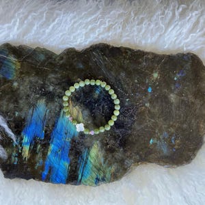 May include: A beaded bracelet with light green beads, small pink beads, and a white flower charm. The bracelet rests on a dark, textured stone with blue and green iridescent patterns. The background is a soft, white, fluffy material.