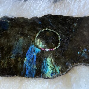 May include: A beaded bracelet featuring green, pink, and white gemstones. The bracelet is displayed on a dark, textured stone with blue and green iridescent patterns. The background is a soft, white, fluffy material.