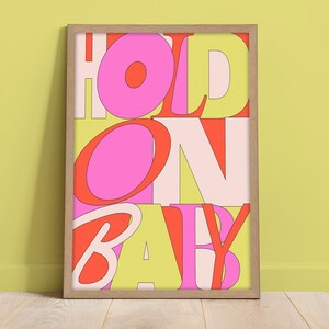 以下が含まれることがあります： 「HOLD ON BABY」というテキストが重なり合ったカラフルな文字で書かれた額装されたプリント。文字はピンク、赤、黄色の色合いです。