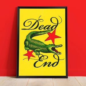May include: A framed art print with a yellow background features a green alligator with an open mouth and two red stars. The words "Dead End" are written in black cursive script above and below the alligator.
