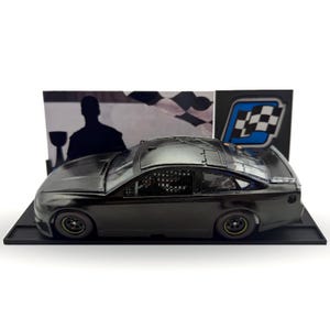 May include: A dark gray die-cast model race car with a black base. The car has a detailed interior and is displayed in front of a backdrop with a checkered flag and a silhouette of a person holding a trophy.
