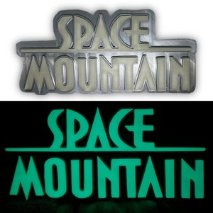 Puede incluir: Un primer plano de un letrero de metal plateado que dice "SPACE MOUNTAIN" en una fuente estilizada. El letrero brilla en verde en la oscuridad.