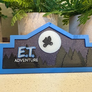 May include: A blue and black sign with the text "E.T. Adventure" and a silhouette of E.T. flying in a spaceship against a white moon.