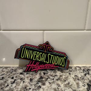 Puede incluir: Un letrero negro y verde de Universal Studios Hollywood con luces de neón. El letrero está sobre una encimera blanca y gris.