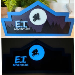 May include: A blue and black sign with the words "E.T. Adventure" and a silhouette of E.T. flying in a spaceship in front of a full moon. The sign glows blue in the dark.