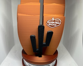 Könnte beinhalten: Eine kupferfarbene Trophäe mit einem Kaktus-Design und einem weißen Band mit dem Text "GEICO" und "Soda Cola". Die Trophäe hat auch einen weißen Aufkleber mit dem Text "Phoenix Raceway Season Finale 500".