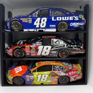 May include: Three NASCAR race car models displayed on black shelves. The top car is blue with the number 48 and the Lowe's logo. The middle car is black with the number 18. The bottom car is multi-colored with the number 18.
