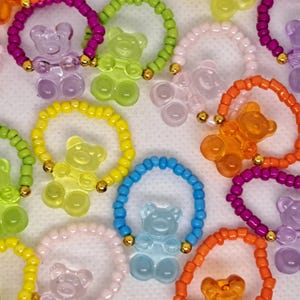 May include: A collection of colorful beaded rings, each featuring a translucent gummy bear charm. The rings come in various colors, including purple, green, yellow, blue, orange, and pink, with gold-colored accents.