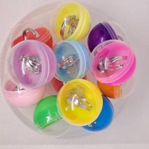 May include: A collection of colorful plastic capsules, each containing a silver-colored ring. The capsules are in various colors, including pink, yellow, blue, and green. The rings have different designs, such as stars and hearts.