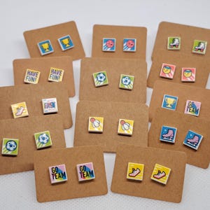 May include: A collection of square stud earrings with sports and activity-themed designs. Each pair is mounted on a small, brown card. Designs include soccer balls, trophies, roller skates, and the words "Have Fun" and "Go Team".