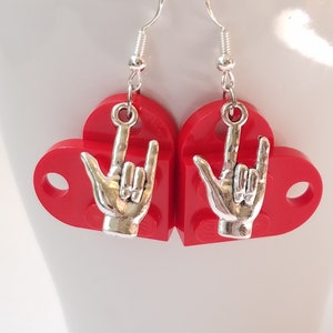 May include: A pair of red heart-shaped earrings with silver-colored hand charms. The earrings have silver-colored hooks for pierced ears. The charms are in the shape of a hand making the 'I love you' sign.