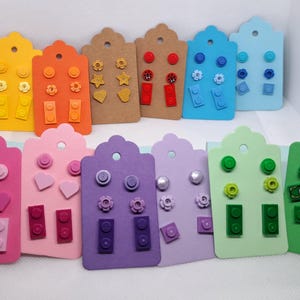 May include: A collection of colorful stud earrings displayed on decorative cards. The earrings feature various designs, including flowers, hearts, and building blocks. The cards are in shades of yellow, orange, brown, blue, pink, purple, and green.