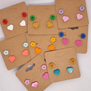 May include: A collection of colorful stud earrings displayed on brown cardboard earring cards. The earrings feature various shapes, including hearts and flowers, in shades of red, white, yellow, green, orange, pink, blue, and teal.