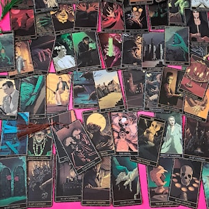 May include: A collection of tarot cards with gothic illustrations, featuring images of skeletons, monsters, and other dark figures. The cards are arranged on a pink surface, with a tassel bookmark visible. The cards have various titles, including "The Moon" and "The Devil".