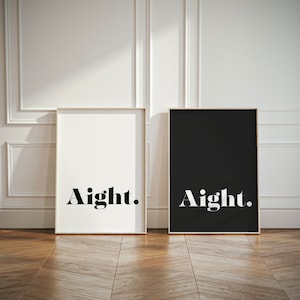 May include: Two framed art prints, one white and one black, lean against a white wall. Each print features the word "Aight." in a bold, serif font. The frames are a light wood color, and the floor is a warm brown.