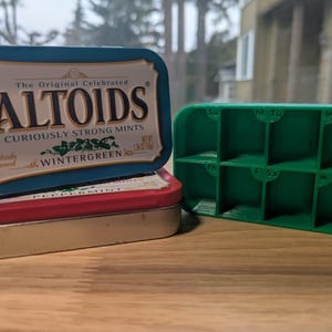 May include: A stack of three containers: a blue Altoids tin, a red and gold tin, and a green pill organizer. The Altoids tin reads "Curiously Strong Mints Wintergreen." The pill organizer is labeled with days of the week.