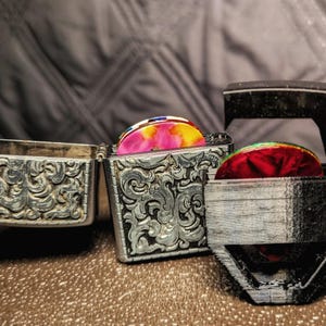 May include: A silver-toned metal case with an intricate embossed scrollwork pattern, shown open with a colorful pink and yellow guitar pick inside, positioned next to a black 3D-printed holder containing a red and green patterned guitar pick.