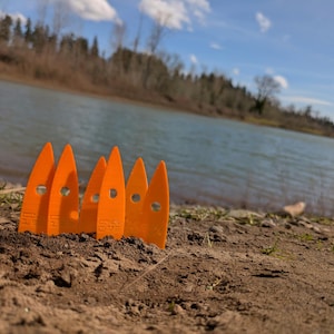 May include: Five orange plastic tent stakes are arranged in a row on a sandy riverbank. The stakes are pointed at the top and have a flat base. The river is in the background.