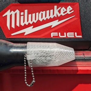 May include: A close-up of a clear plastic nozzle with a chain attached. The nozzle is attached to a black tool. The tool is sitting on top of a red toolbox with the Milwaukee FUEL logo.