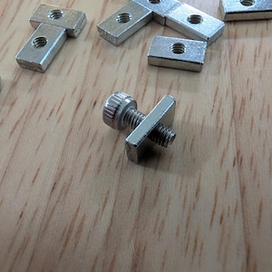 May include: A collection of small, silver-colored metal T-nuts and a bolt with a square head. The T-nuts are rectangular with a threaded hole in the center. The bolt has a knurled head and a threaded shaft. The items are on a light brown wooden surface.