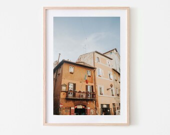 May include: A framed photograph of a building in a city. The building is a light orange color with a balcony and a flag hanging from it. The building is next to a taller building that is a light peach color. The sky is a light blue color.