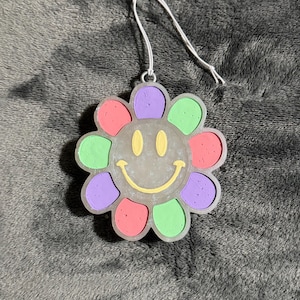 May include: A colorful flower-shaped ornament with a smiley face in the center. The petals are in shades of pink, green, and purple. A silver wire loop is attached for hanging. The ornament is likely made of resin or a similar material.