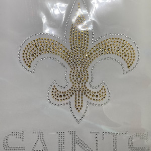 May include: A white fabric with a gold and silver rhinestone fleur-de-lis design and the word "SAINTS" in silver rhinestones.