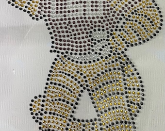 May include: A large, cartoon-style mascot made of rhinestones. The mascot is wearing a maroon jersey and gold pants. The mascot is holding a football in its right hand and is in a running pose. The mascot is made of black, gold, silver, and maroon rhinestones.
