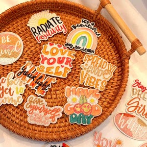 Könnte beinhalten: Eine Sammlung von gestickten Aufnähern mit positiven Affirmationen und farbenfrohen Designs. Die Aufnäher enthalten Sätze wie „Radiate Positivity“, „Love Yourself“ und „Spread Good Vibes“. Sie sind auf einem gewebten Weidenkorb mit Holzgriff angeordnet.