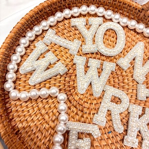 Könnte beinhalten: Nahaufnahme von dekorativen, mit Perlen bedeckten Buchstaben und einer Perlenkette, die auf einem Weidenkorb angeordnet sind. Die Buchstaben sind weiß mit einem Perlen- und Strassdesign. Die Perlen sind weiß.